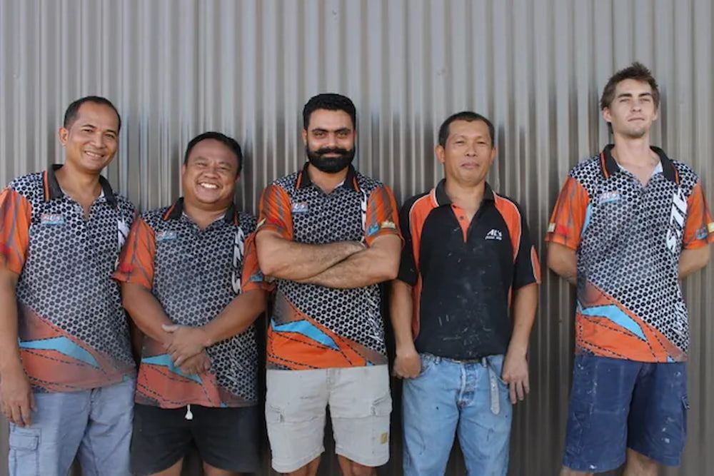 Five Men in Matching Orange and Black Shirts Stand in Front of A Corrugated Wall — Al's Panel Shop in Berrimah, NT