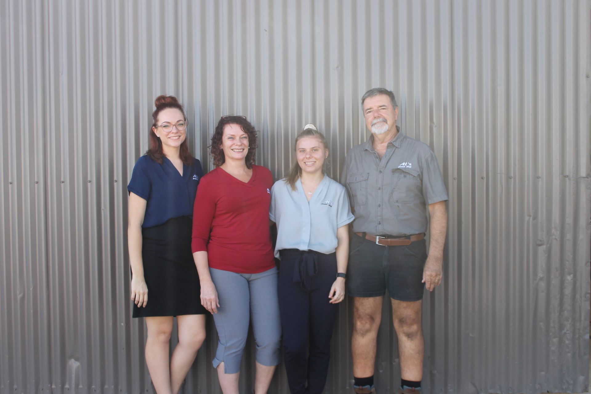 Four people stand smiling against a gray corrugated metal wall, wearing casual tops, shorts, skirts, and trousers.