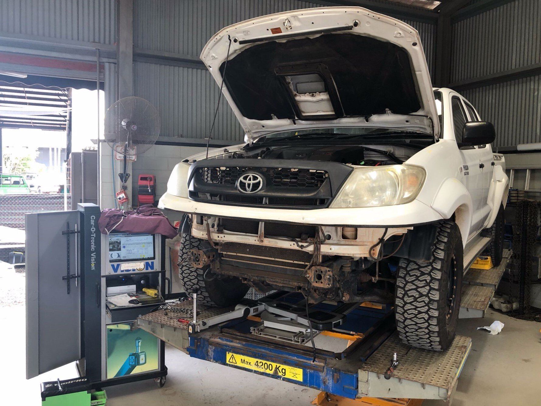 A white Toyota Hilux truck with its hood open is elevated on a lift in a mechanical workshop.
