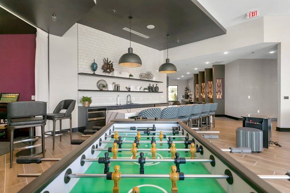 Modern lounge with foosball table, bar seating, and wall shelves under pendant lights at Haven on Tucker Apartments in Louisville, KY.