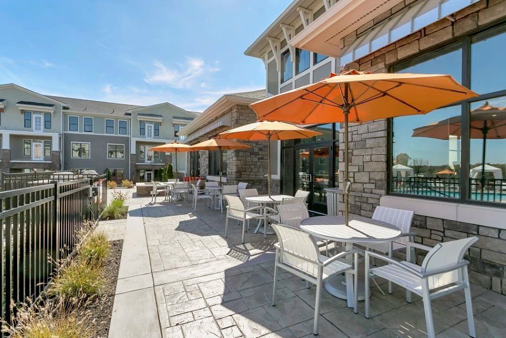 Outdoor patio seating with orange umbrellas and white tables beside apartment buildings at Haven on Tucker Apartments in Louisville, KY.