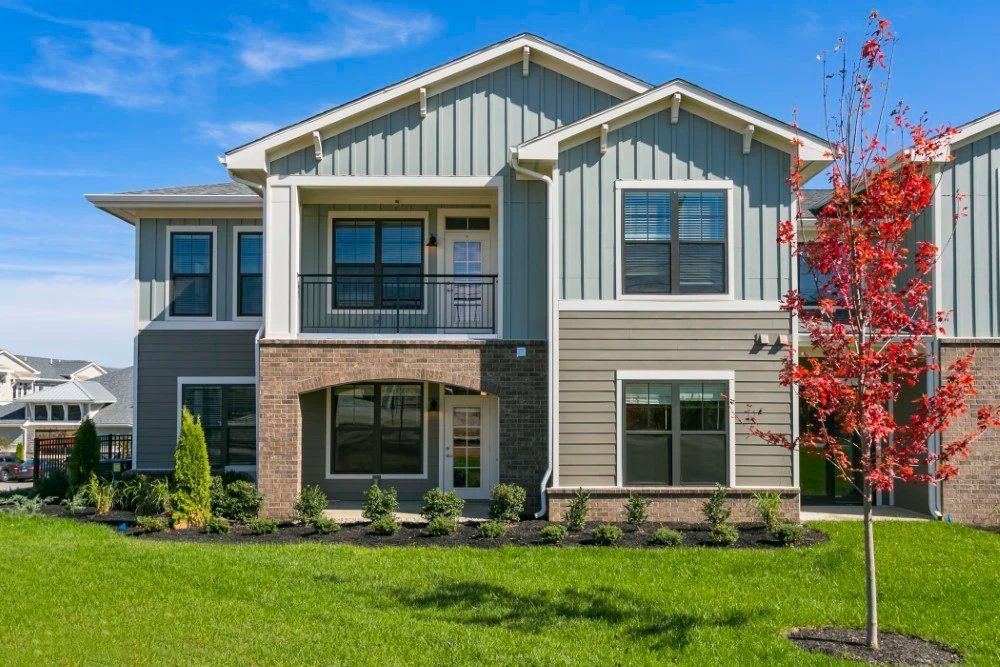 Modern two-story suburban house with gray siding, stone facade, green lawn, and a red-leafed tree under a blue sky at Haven on Tucker Apartments in Louisville, KY.