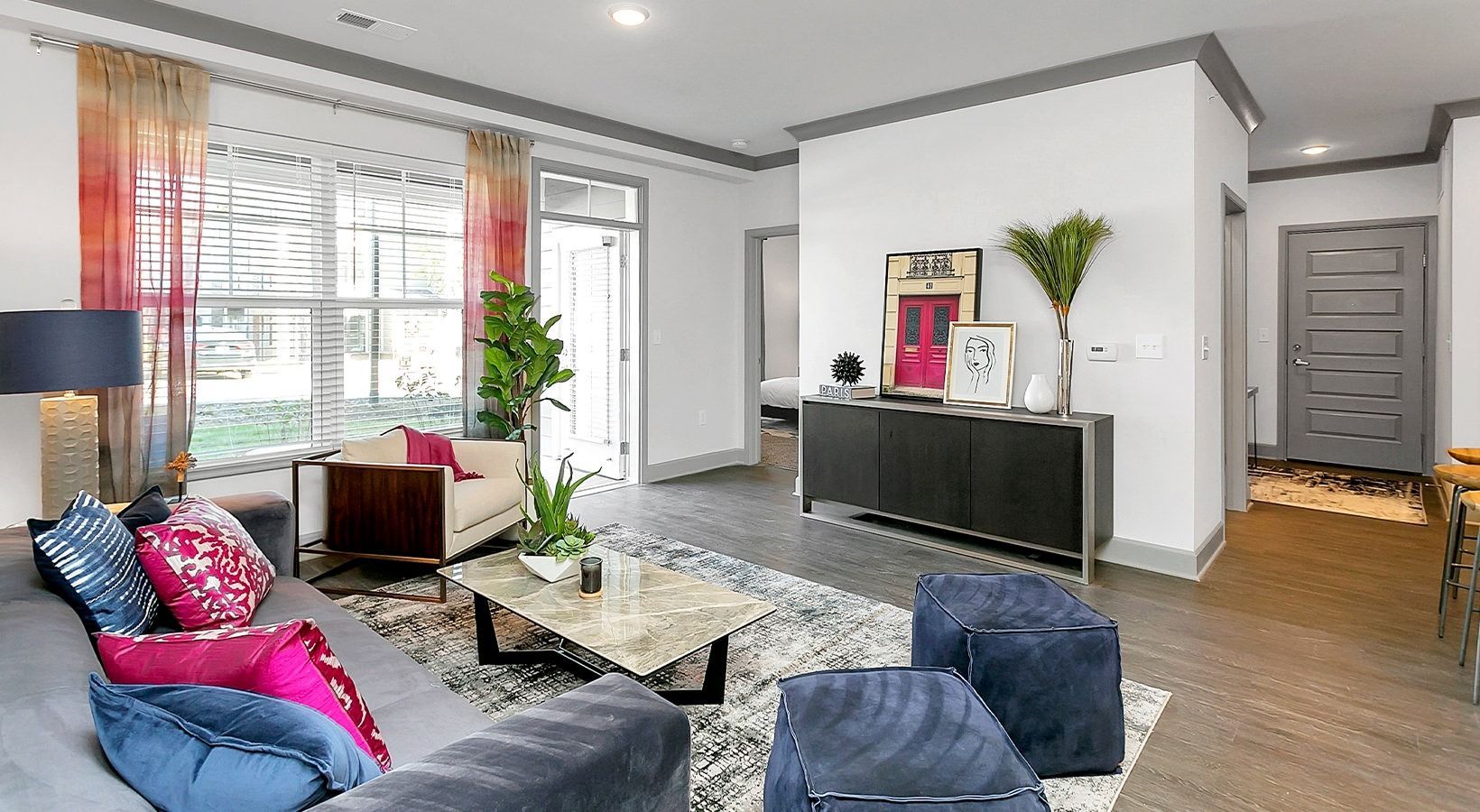 Bright modern living room with blue sofas, colorful cushions, glass coffee table, and large windows at Haven on Tucker Apartments in Louisville, KY