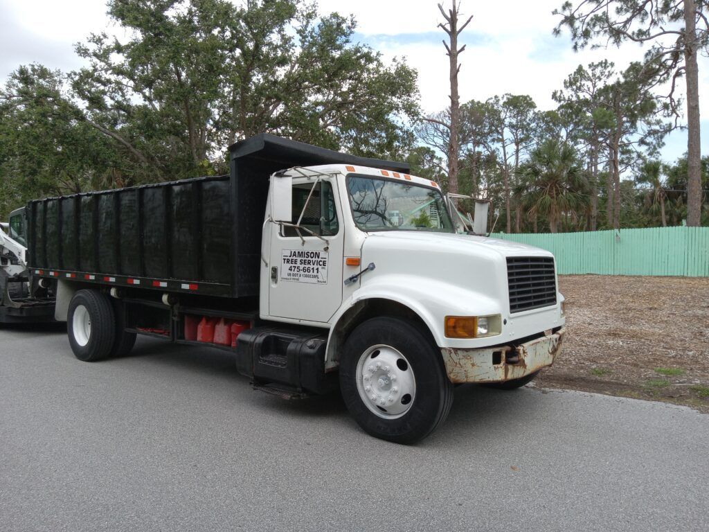 White dump truck parked on a road beside trees and a green fence