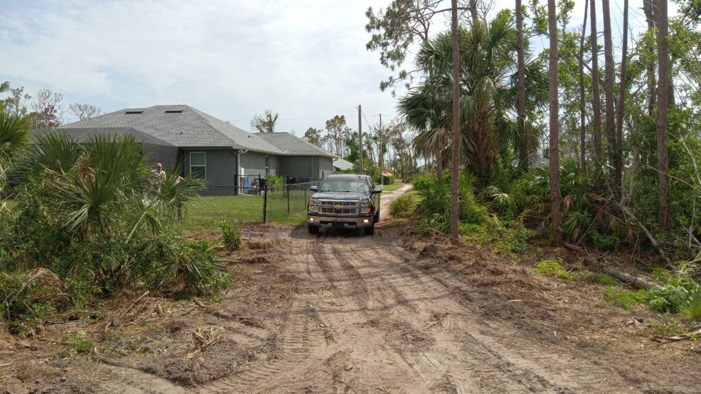 Truck on a muddy dirt driveway beside a house and tall trees.