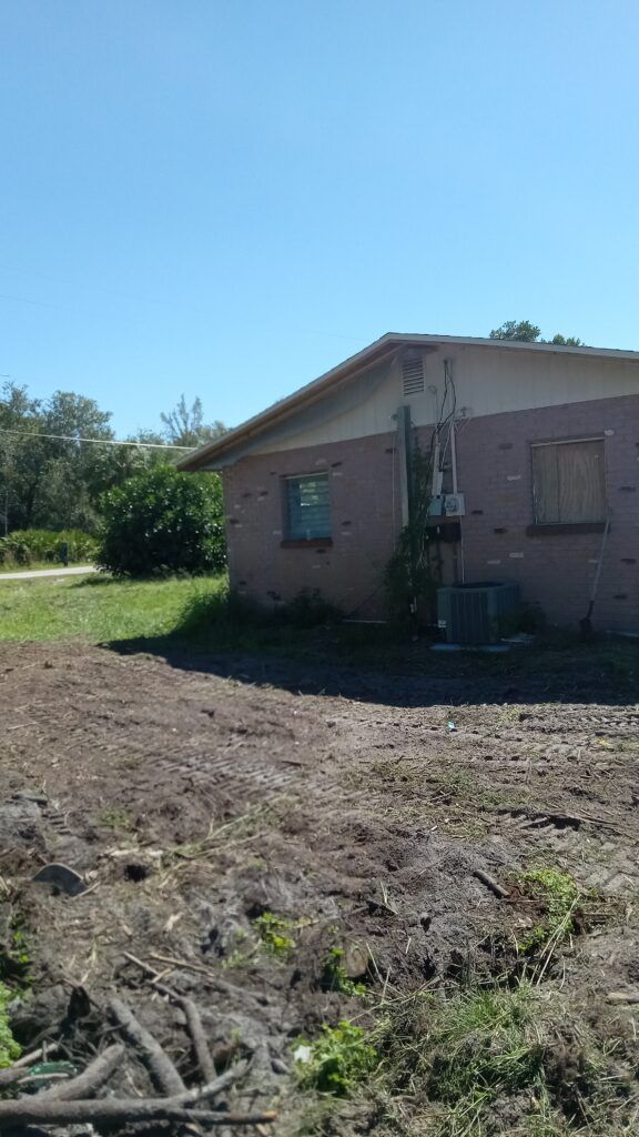 Small brick house with boarded windows in a muddy yard under a clear blue sky