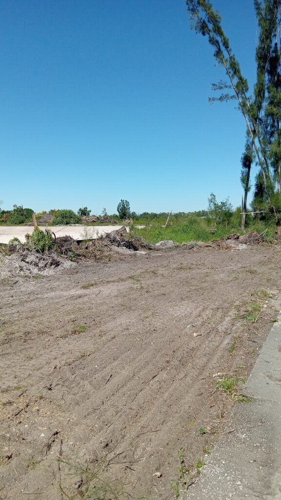 Cleared dirt lot with a mound of rubble beside a paved road under a clear blue sky