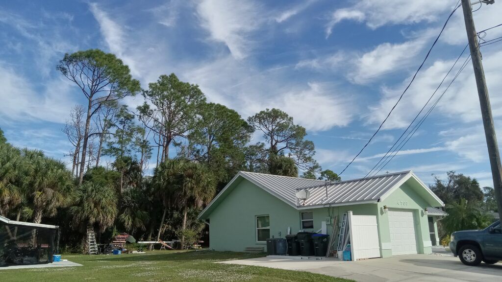 Small mint-green house with a metal roof beside a road, under a blue sky with trees and power lines.