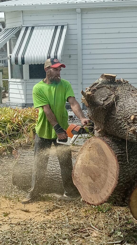 Person in green shirt using a chainsaw to cut a large log outside a house