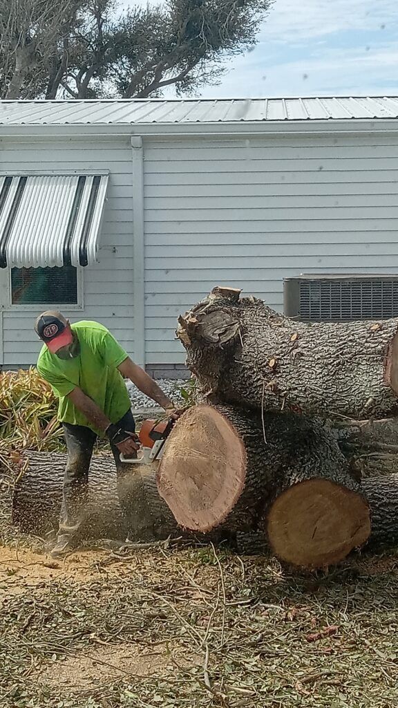 Worker in neon shirt cutting a large log beside stacked timber outside a white house.