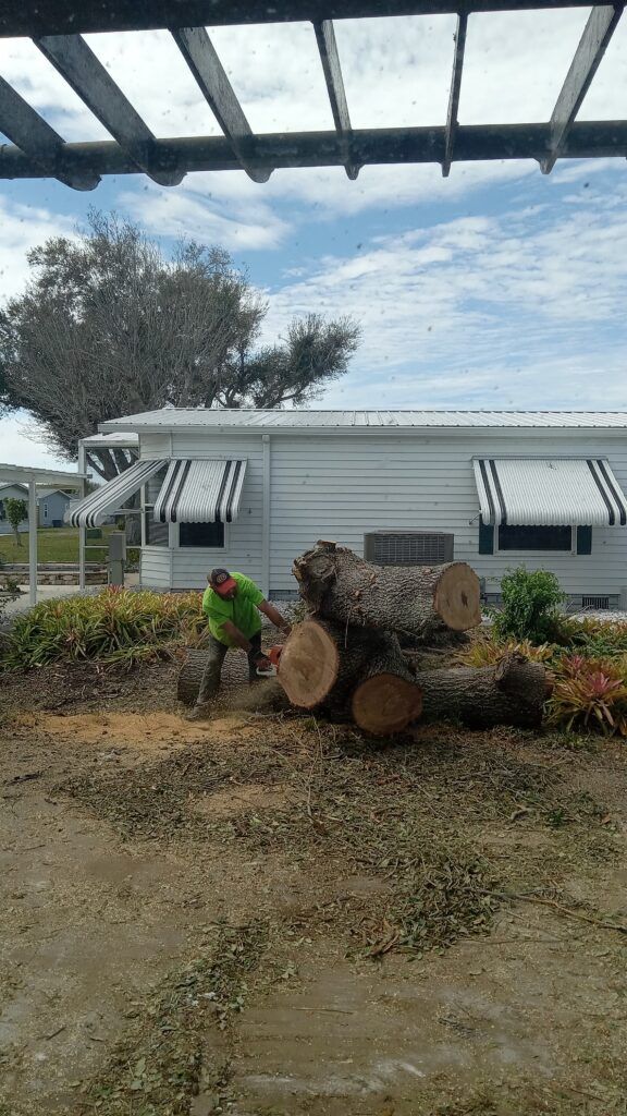 Person in green shirt working beside stacked logs in a yard with a white trailer and awning.