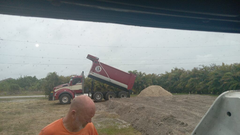 Red dump truck tipping gravel beside a dirt road, with a person in an orange shirt in the foreground.