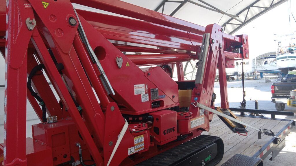 Red construction lift with folded boom parked indoors near a waterfront.