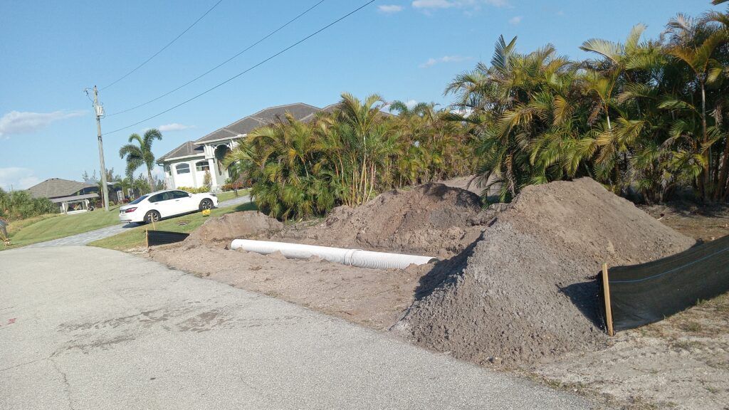 Piles of gravel and dirt beside a residential street with a white truck and palm trees under clear sky