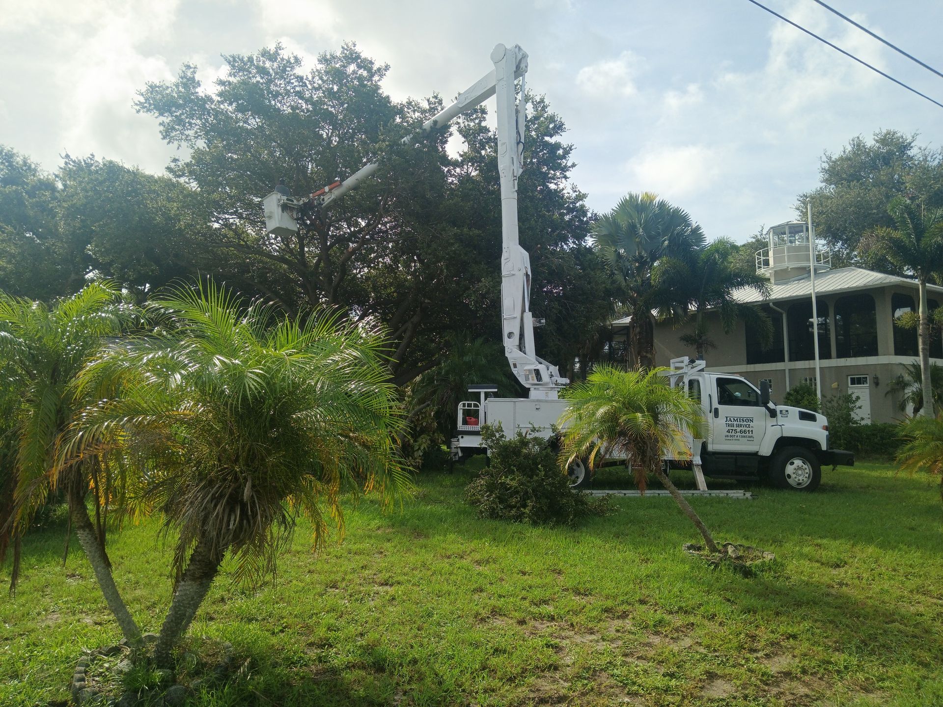 White utility truck with raised boom in a grassy yard near a house and trees, under a cloudy sky