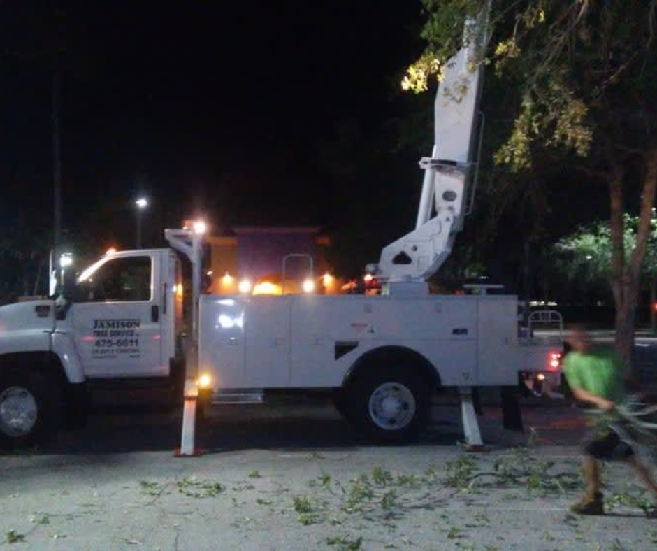 White utility bucket truck with raised boom parked at night on a street, cones around it and a worker nearby