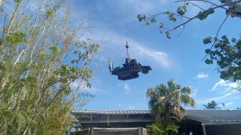 A roller coaster car hangs from an overhead track above trees and a building under a blue sky.