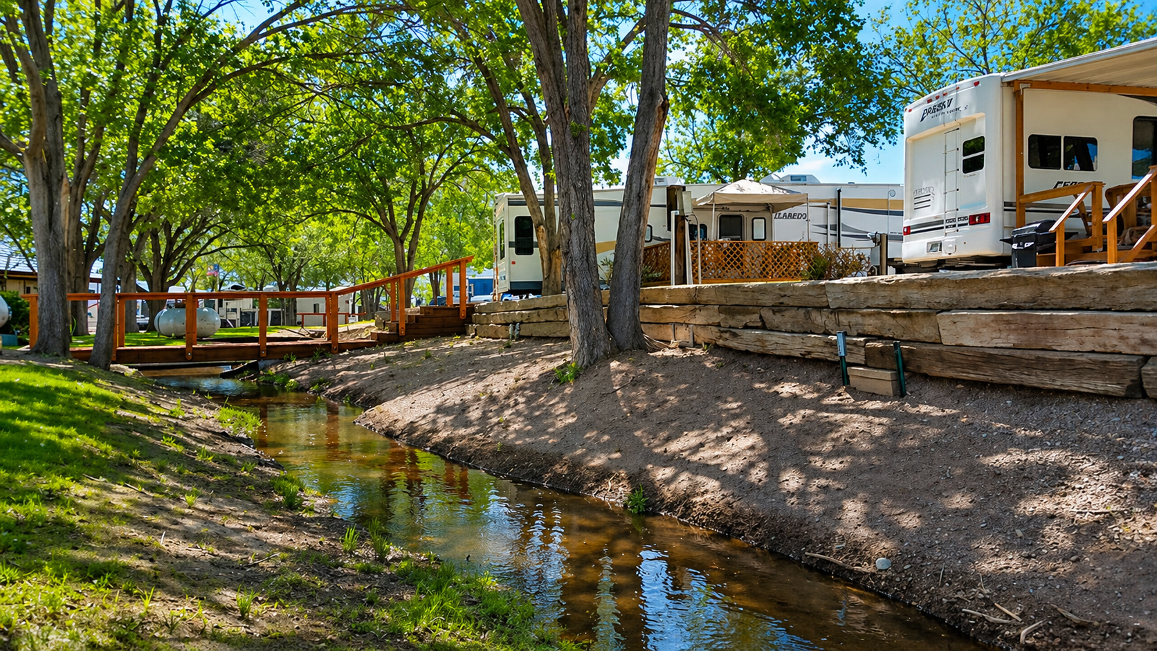 Tree-lined creek beside RVs and a wooden deck in a sunny park.