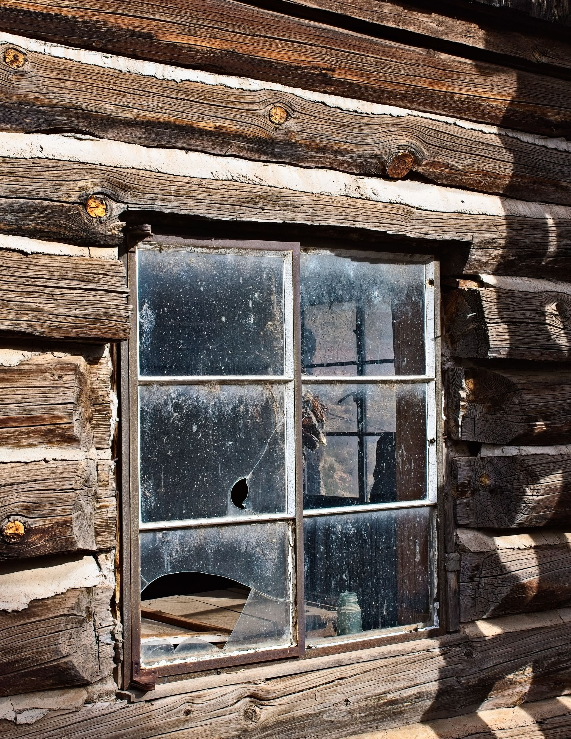 Broken-paned window on a weathered log cabin wall with peeling paint and reflected trees