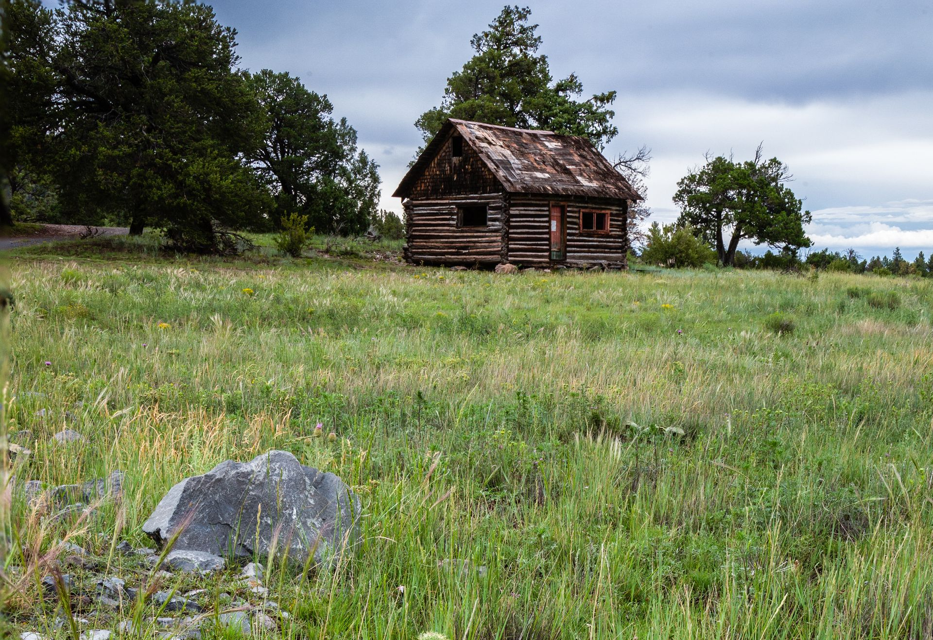 Small wooden cabin in a grassy field with trees and a cloudy sky