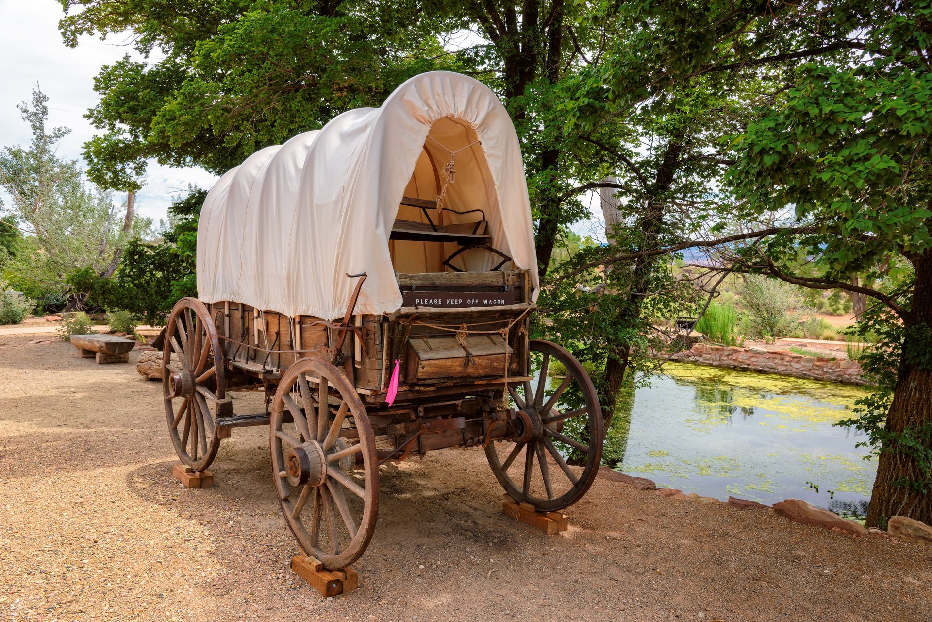 Covered wooden wagon by a river, with large wheels and a white canopy under trees.