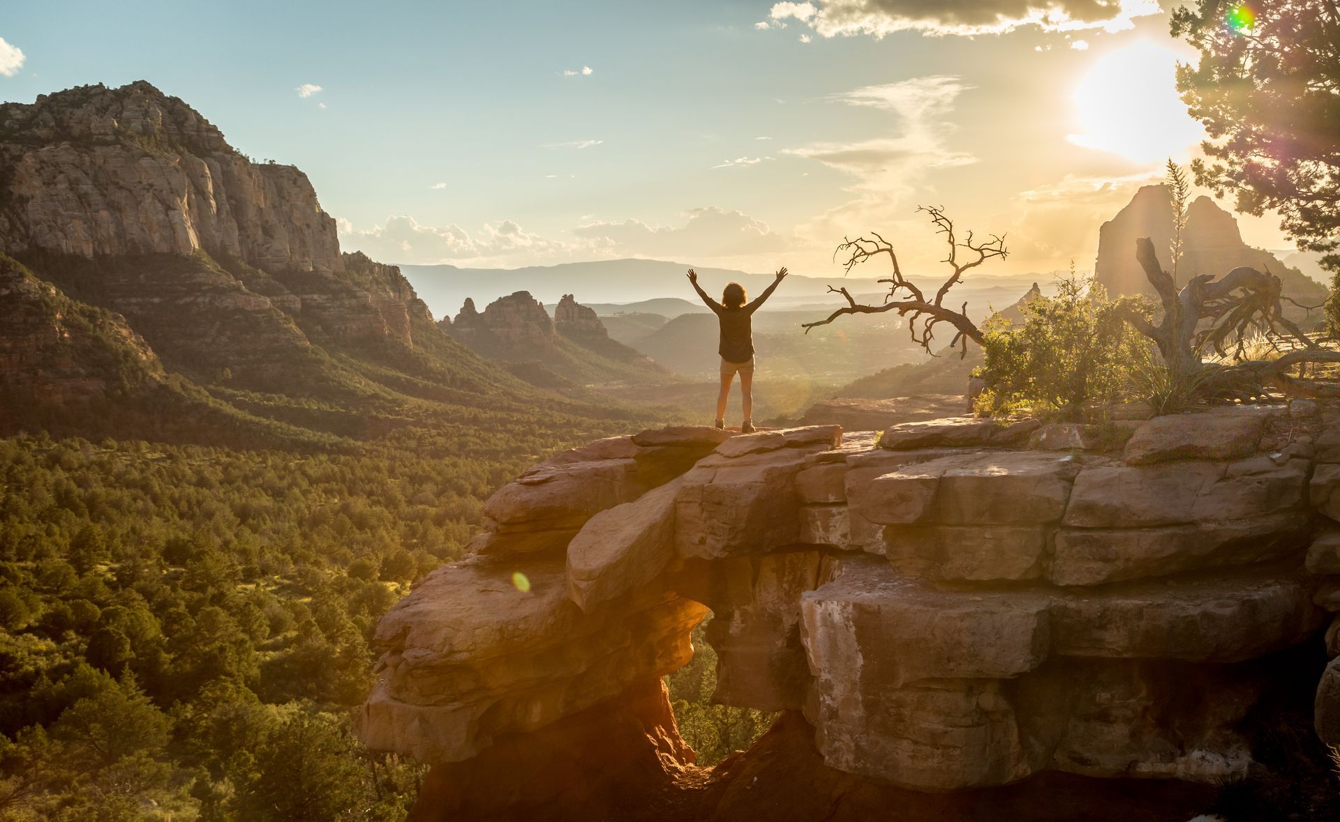 Person standing on a rocky cliff at sunrise with arms raised, overlooking a lush mountain valley