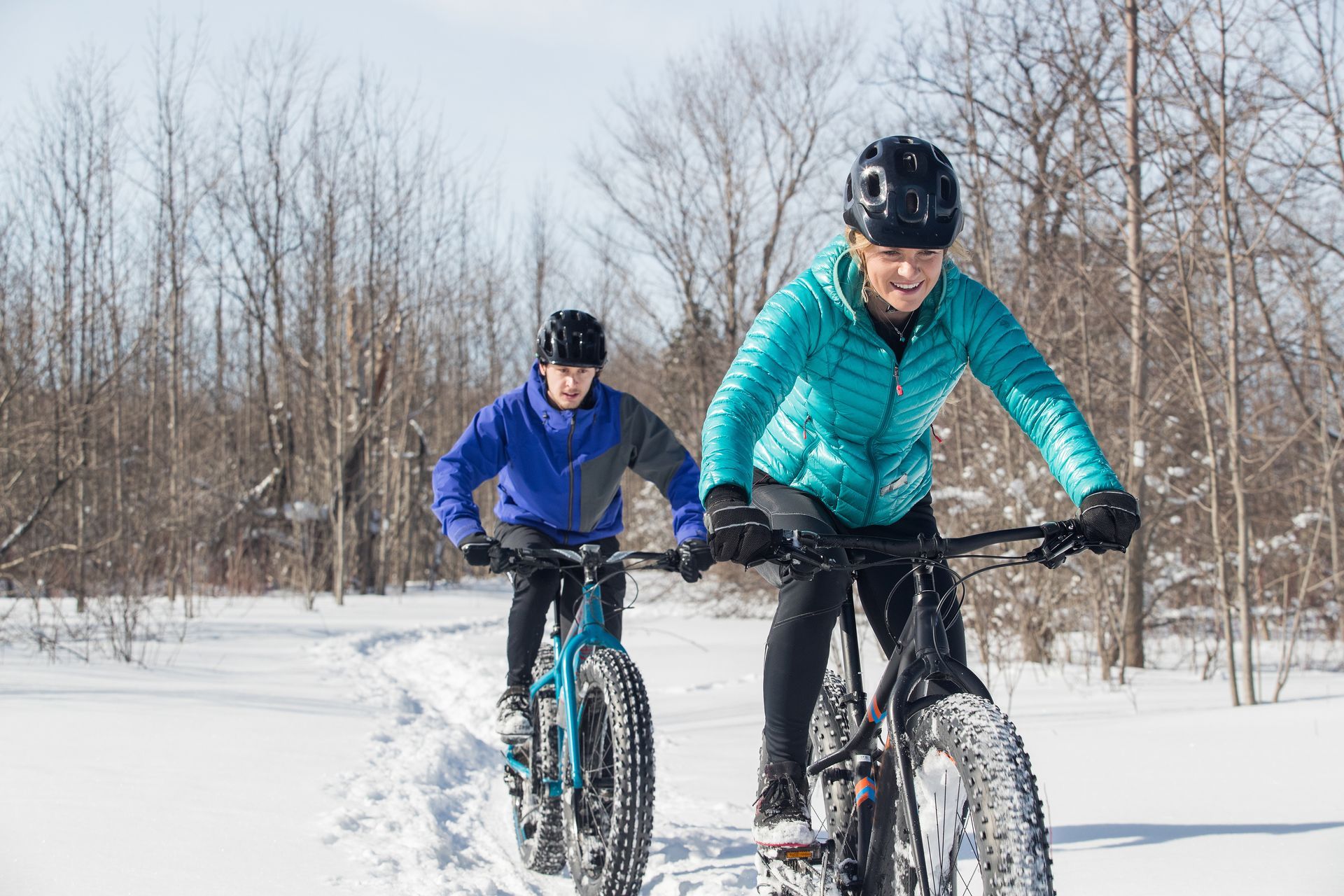 Two cyclists riding fat-tire bikes through snowy woods, one in teal and one in blue.