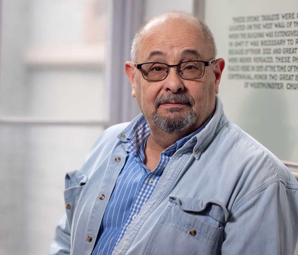 Man with glasses, light blue shirt, and goatee, seated, looking at the camera. White building background.