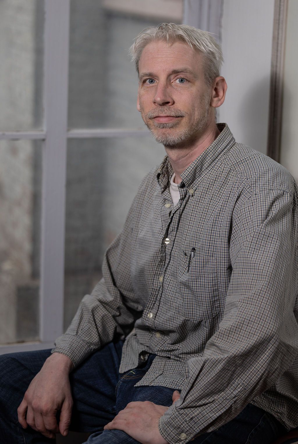 Man with graying hair, seated by a window, wearing patterned shirt, jeans.