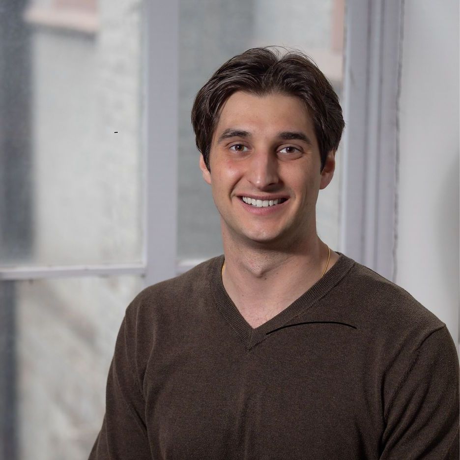 A man in a brown sweater is smiling in front of a window.