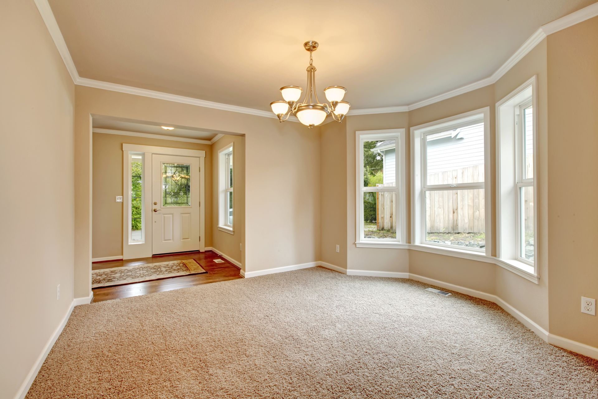 Bright empty living room with beige carpet, bay windows, chandelier, and open doorway to a foyer