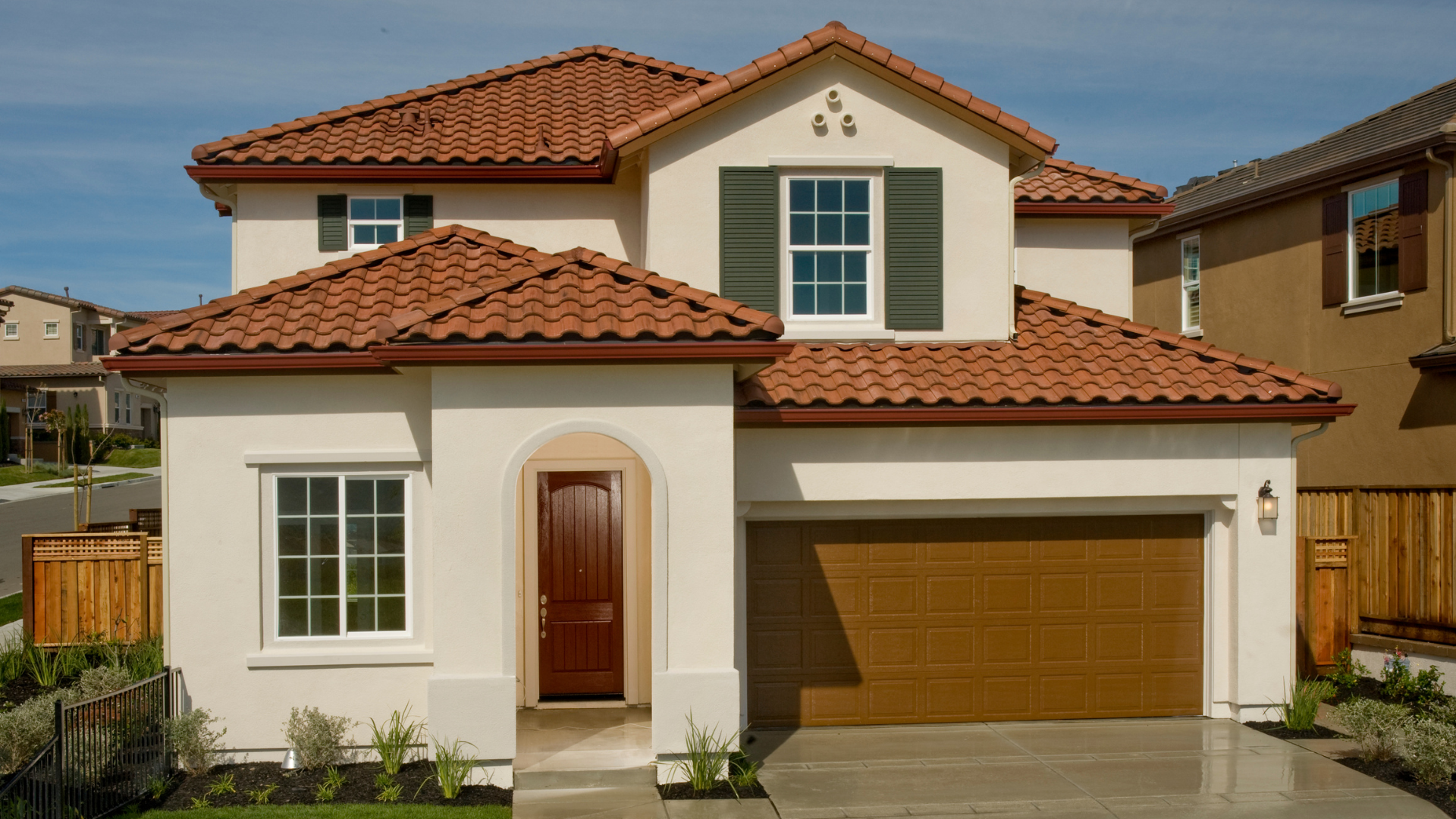 Two-story beige house with red tile roof, white trim, and a brown garage door behind a small front yard