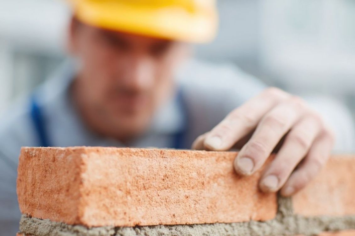A construction worker in a yellow hard hat places a brick on a wall, focus on brick.