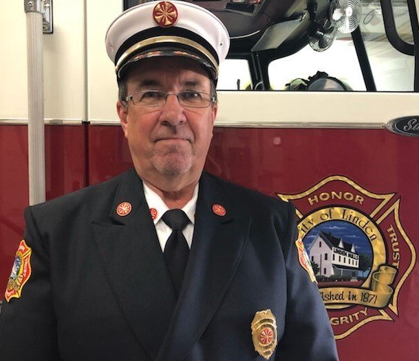 Firefighter in uniform stands in front of fire truck. He wears glasses, a cap, and a badge.