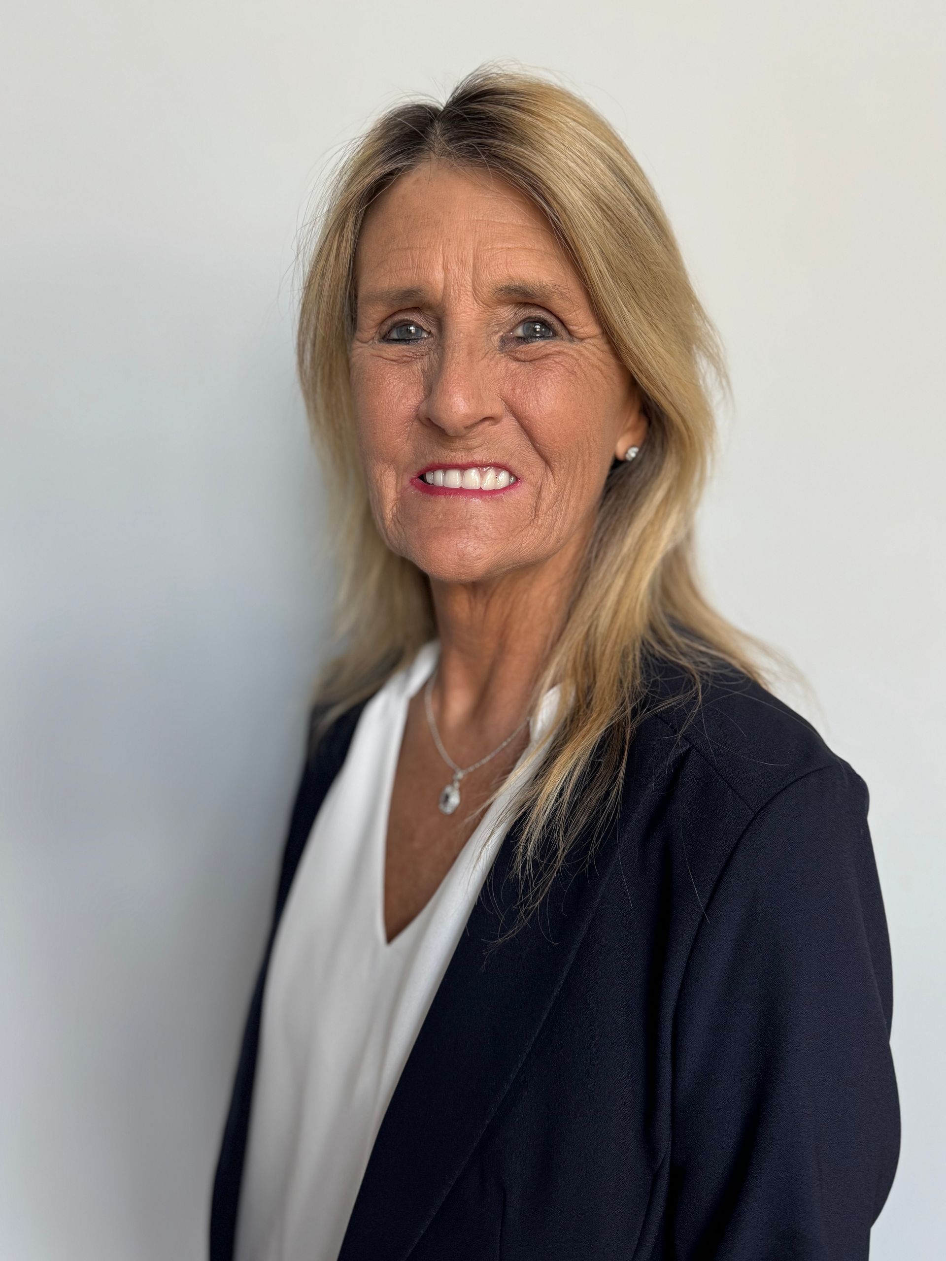 Woman with blonde hair wearing a dark jacket and white shirt smiles, standing against a white wall.