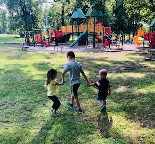 Three children are holding hands in a park in front of a playground.
