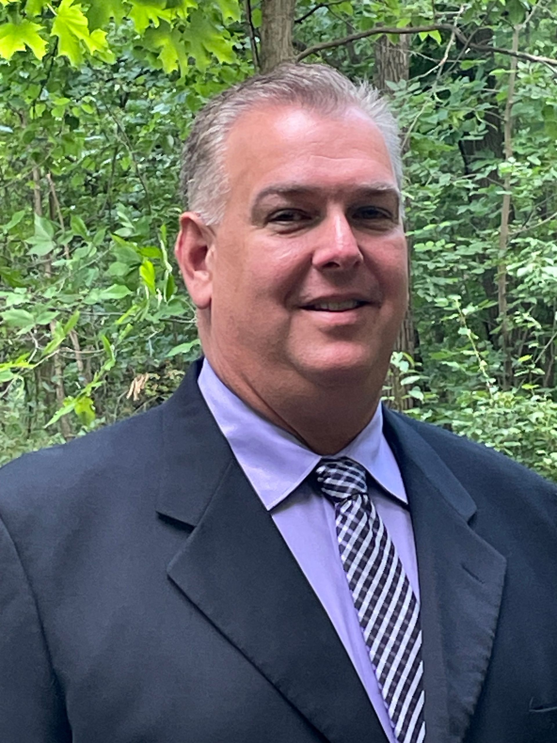 Man in a suit, light purple shirt, and patterned tie smiling outdoors with a green background.