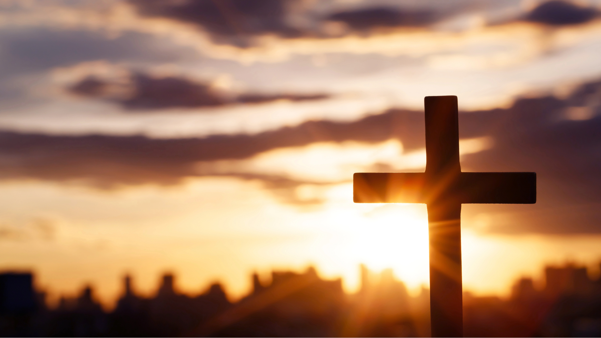 Silhouette of a wooden cross at sunset with a glowing sky and blurred city skyline.