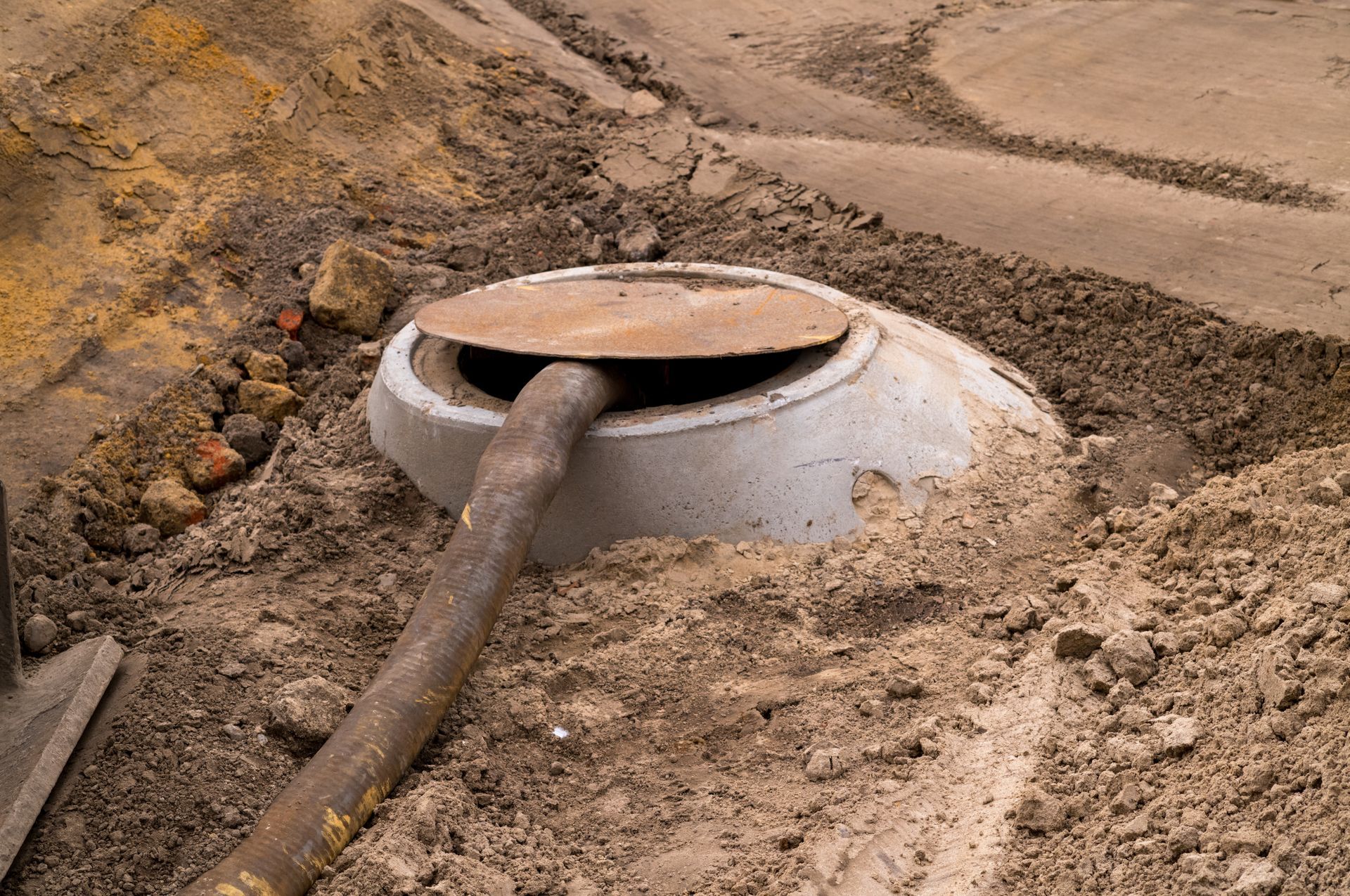 A hose runs into an open concrete manhole in the middle of a dirt excavation site.