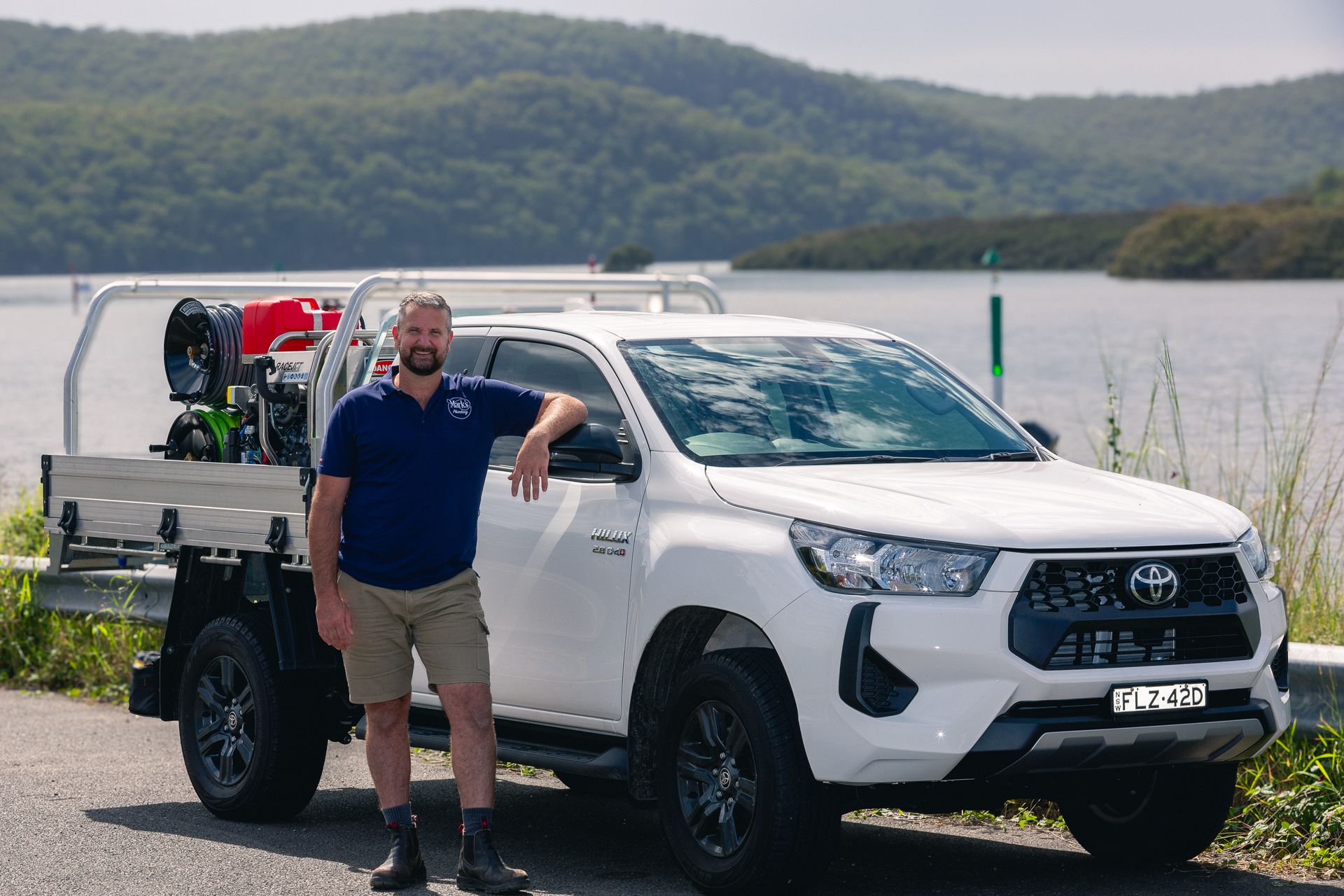 Man Standing Beside a White Toyota Pickup With Equipment, Parked by a Lake and Hills — Mark's Plumbing in Noraville, NSW