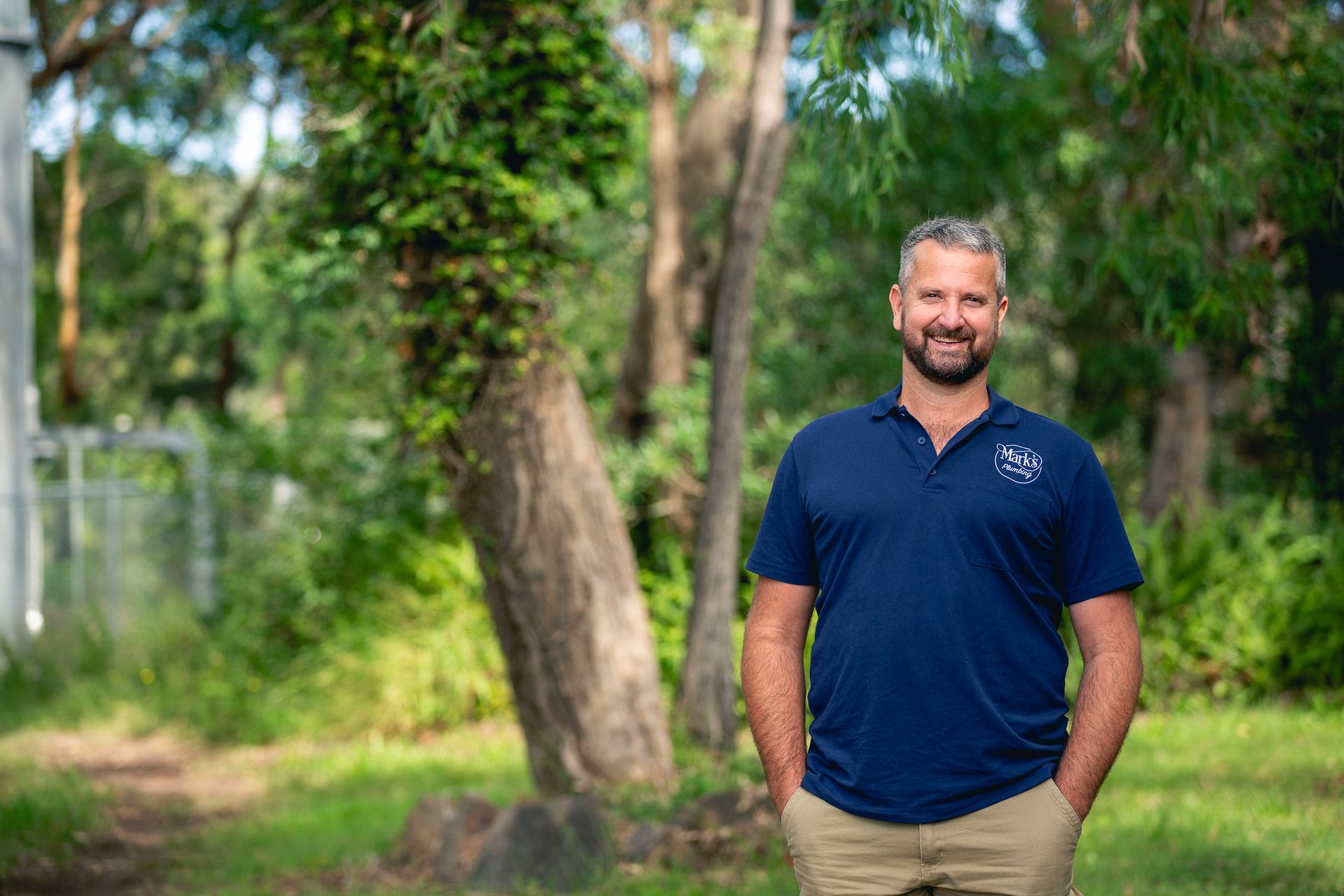 Man Standing Outdoors in a Blue Polo Shirt and Khaki Pants, Smiling — Mark's Plumbing in Noraville, NSW