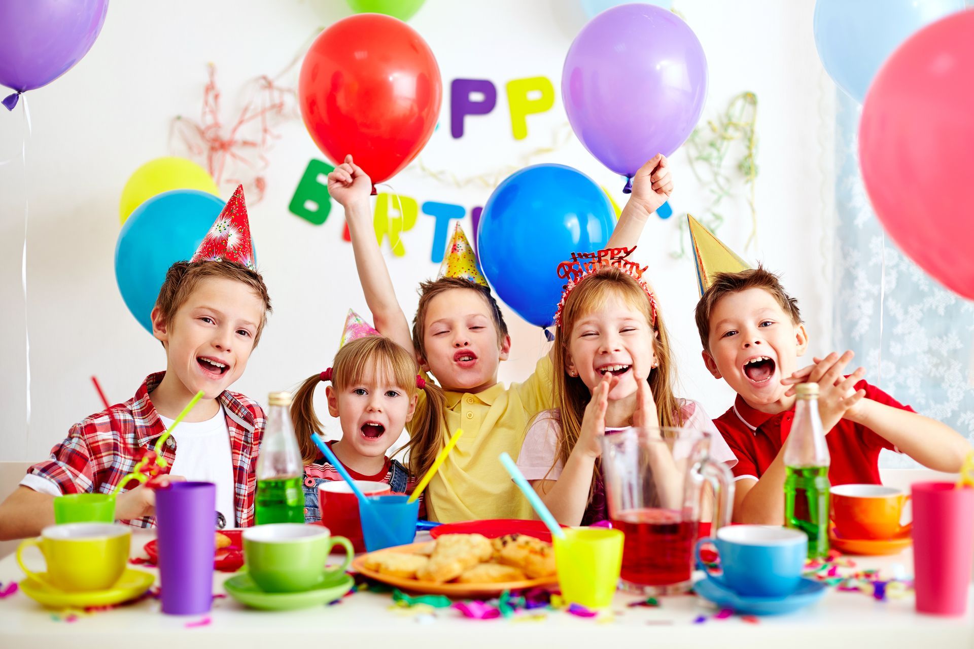 Children celebrating a birthday, raising balloons, and smiling at a decorated table with drinks and treats.
