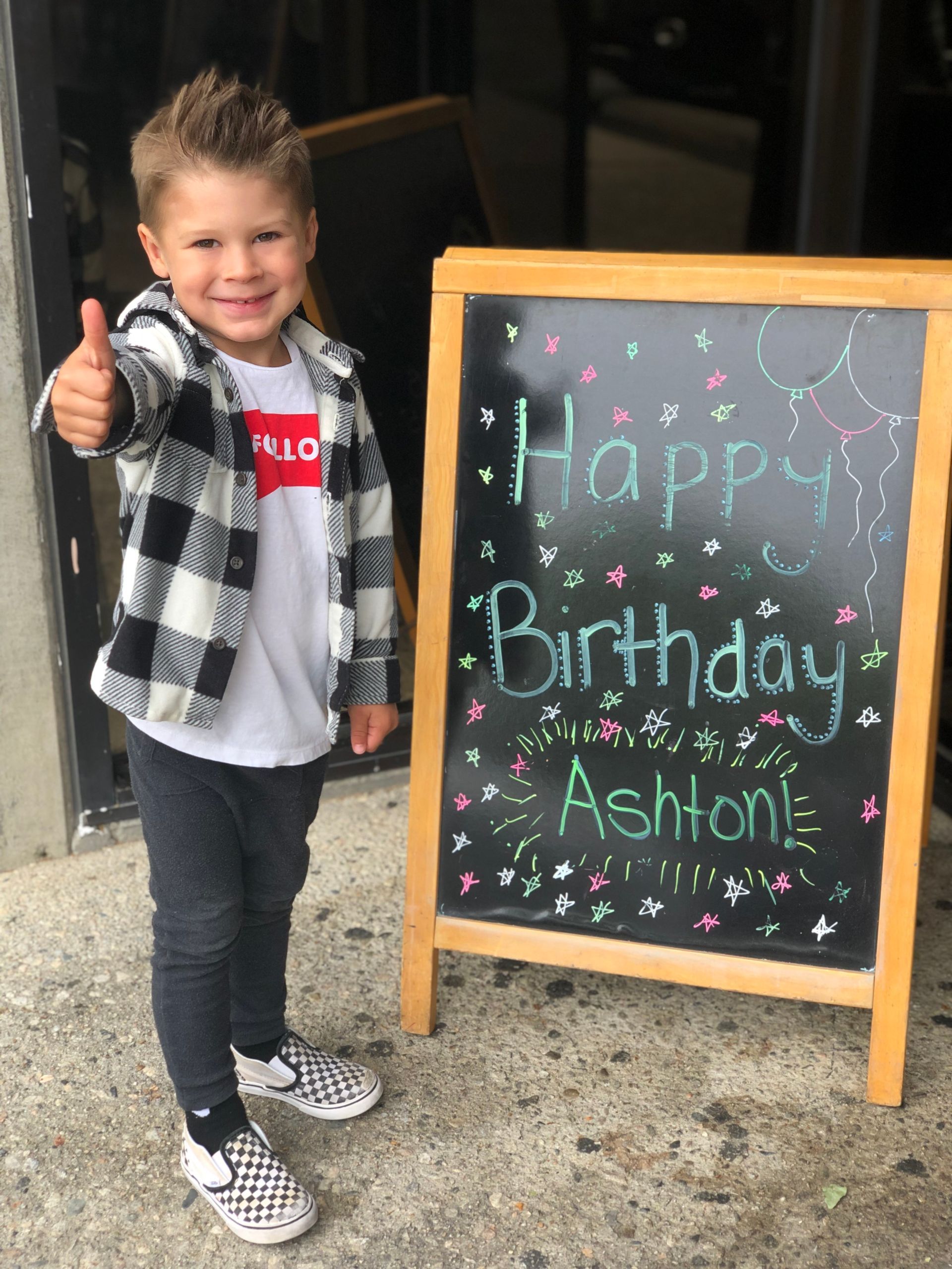 Boy giving thumbs up next to a chalkboard sign that says