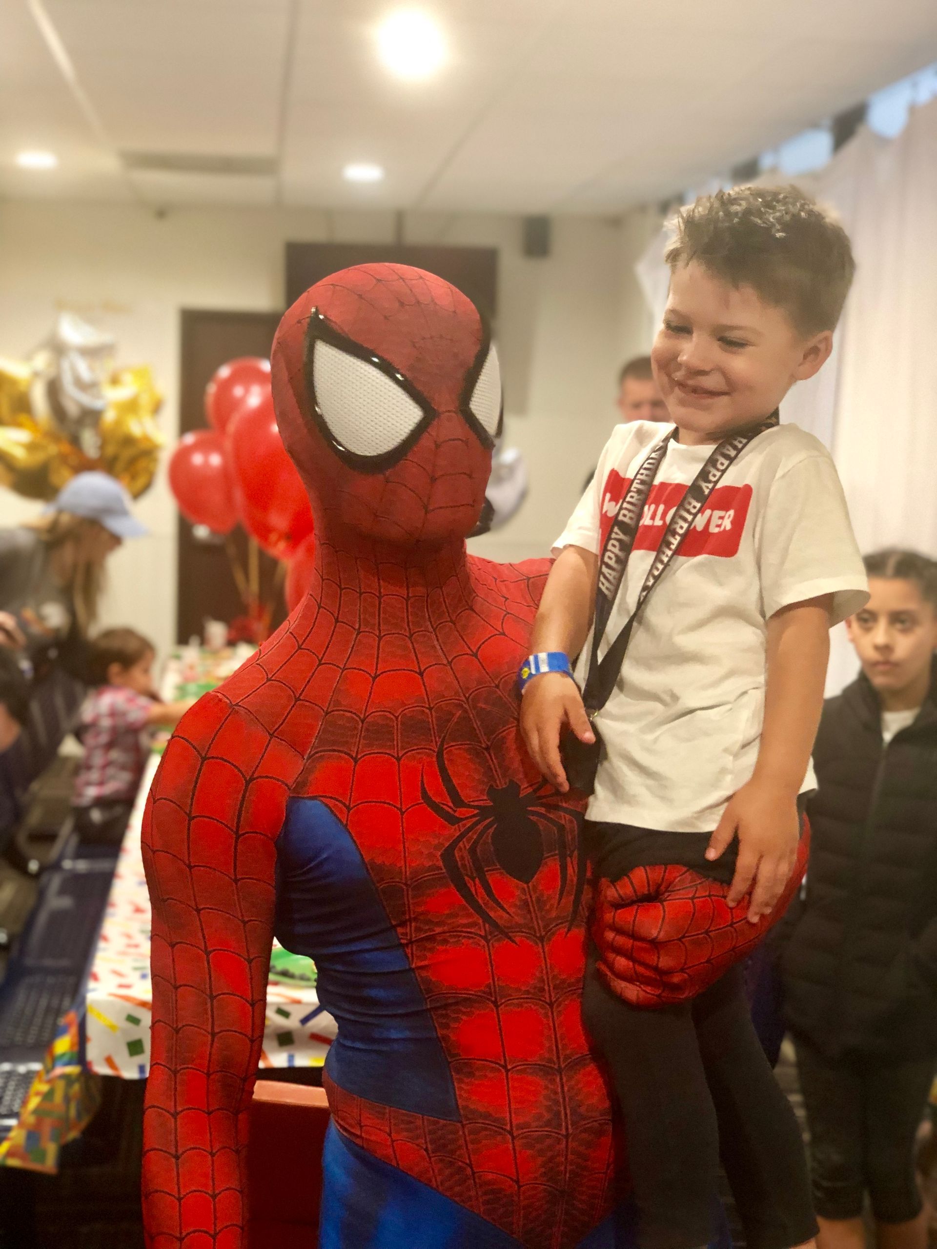 Spider-Man holds a smiling child in a party setting with balloons.