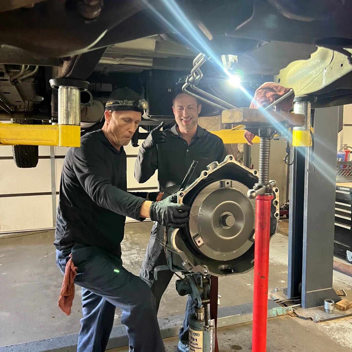 Two workers inspect machinery under a vehicle lift in a garage, with one holding a large circular part. | Flagstaff Auto Repair