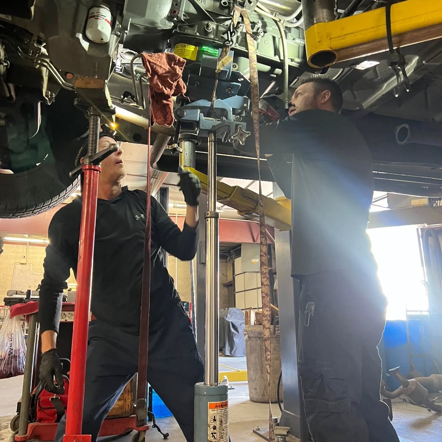 Two mechanics work under a vehicle lift in a busy garage, adjusting parts overhead. | Flagstaff Auto Repair