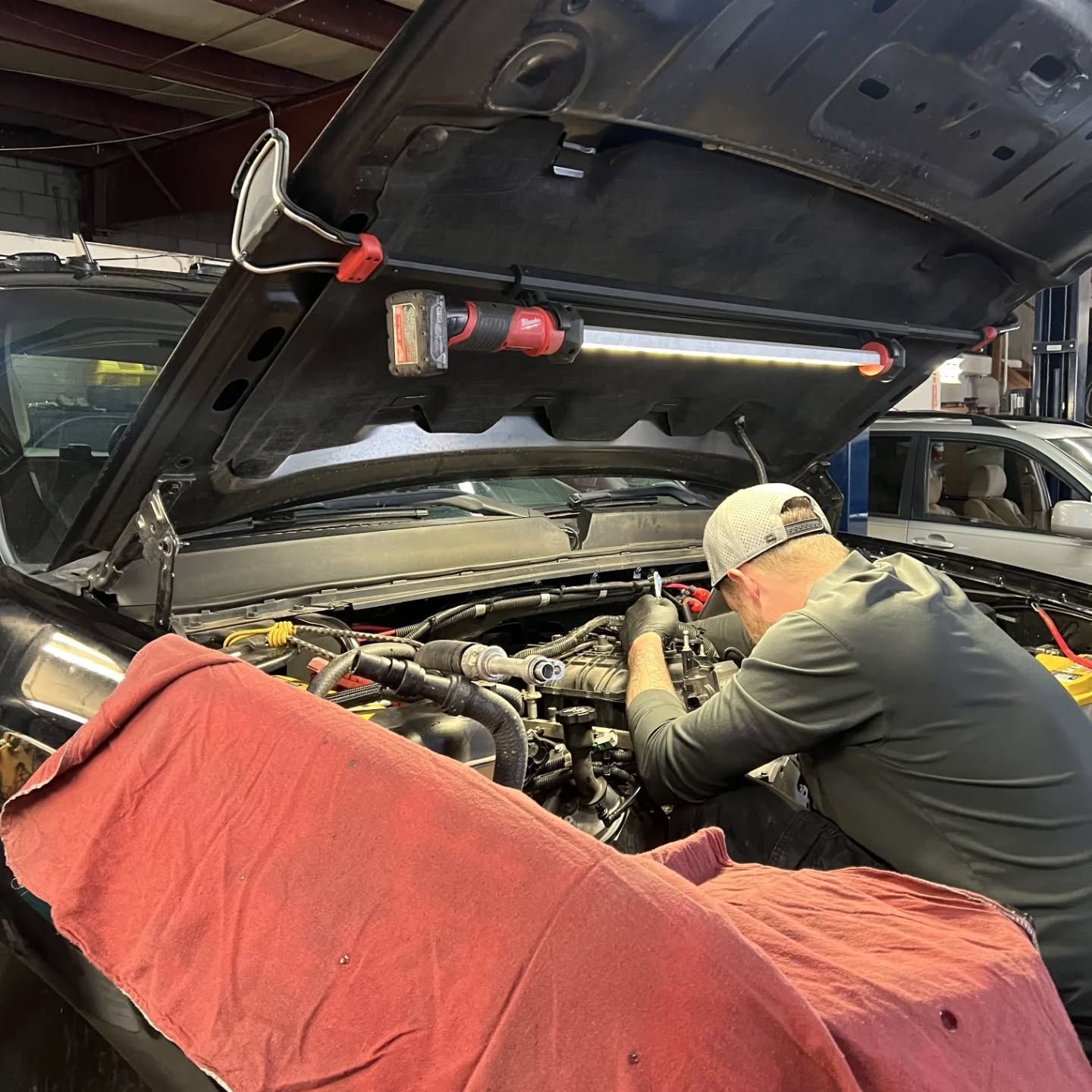 Mechanic working under an open car hood in a garage, with a red fender cover draped over the front. | Flagstaff Auto Repair