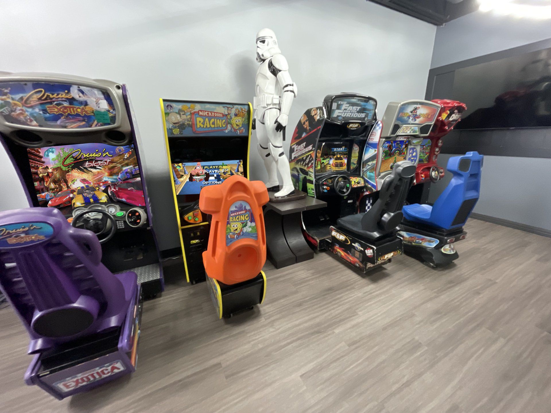 A row of arcade racing games, featuring a stormtrooper statue between them, set in a room with grey walls and wood flooring.