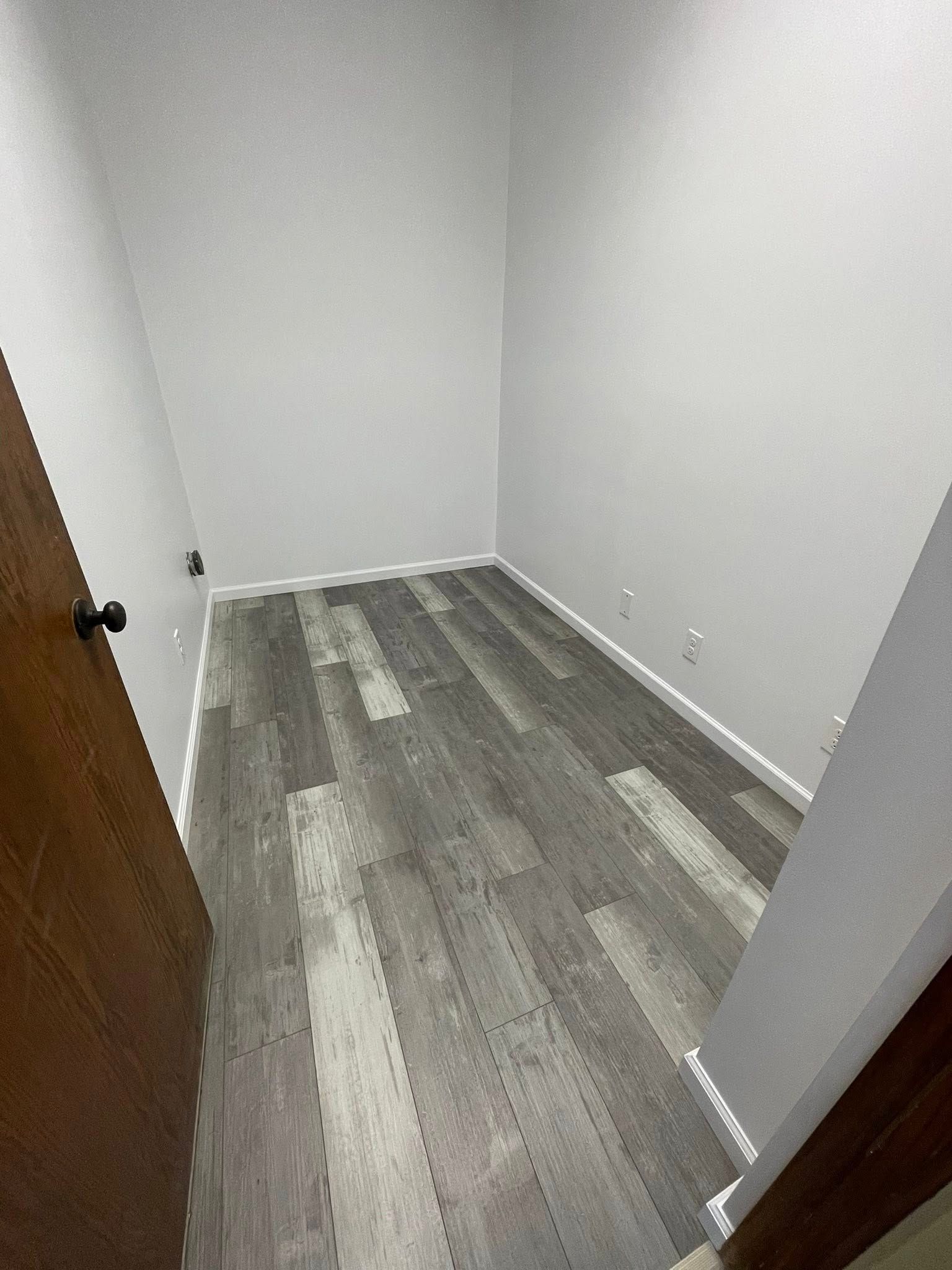 Small, empty closet with gray wood-look flooring, white walls, and a brown door.
