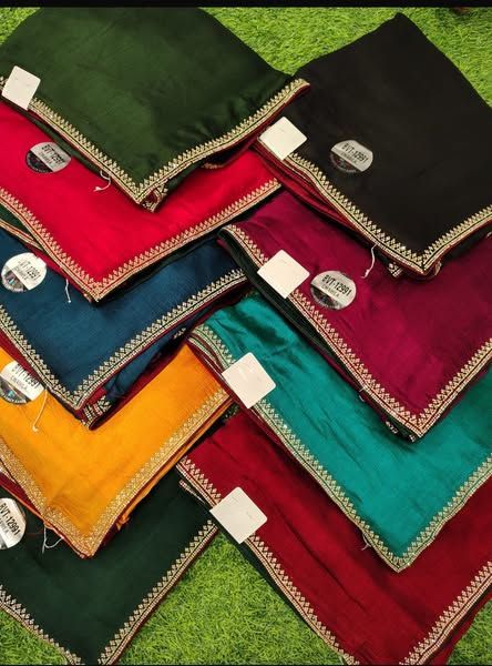 Pile of colorful silk sarees with embroidered edges, arranged on green.